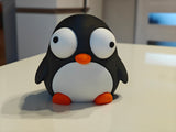 Klein pinguïnfiguurtje met beweegbare ogen op een bureau bij een koffiemok